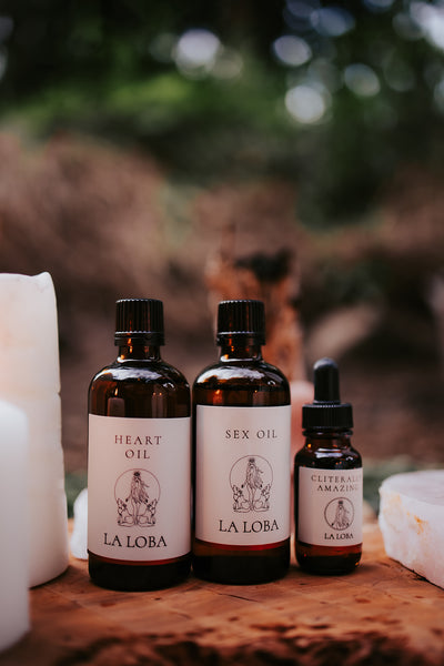 Three bottles of sensory oil products, labeled as 'Heart Oil', 'Sex Oil', and 'Cliterally Amazing Oil', presented on a wooden surface with candles in the background.
